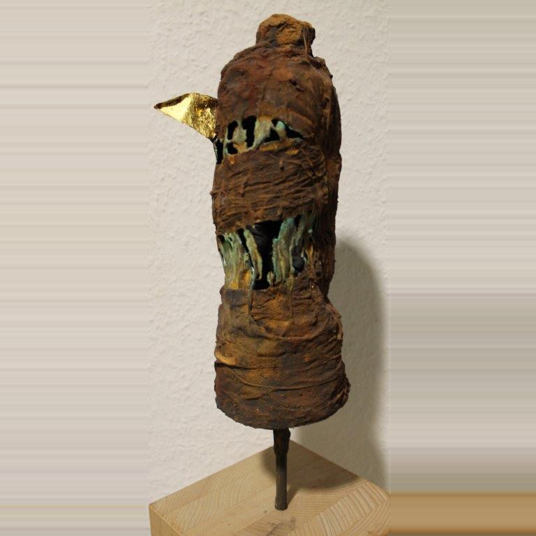 Gedrungene Skulptur in Rostbraun mit grünen Einschlüssen und einer goldenen Spitze, auf einem Stahlstift über einem Holzsockel.