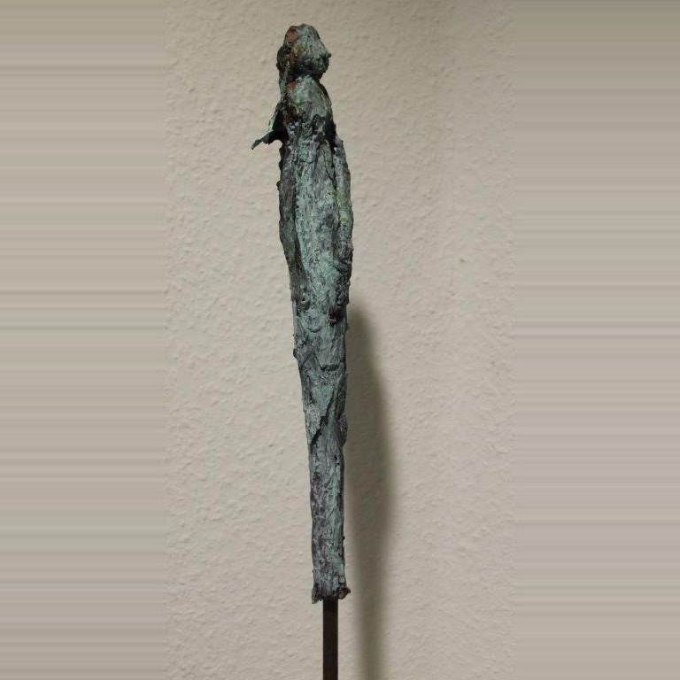 Hoch aufragende, sehr schmale Figur mit grün-blauer Patina, auf einem dünnen Stahlstift vor heller Wand.